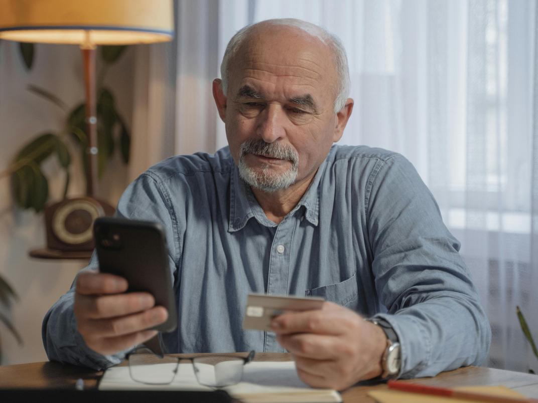 Oudere ma zit aan een tafel en heeft zijn telefoon en een bankpas vast