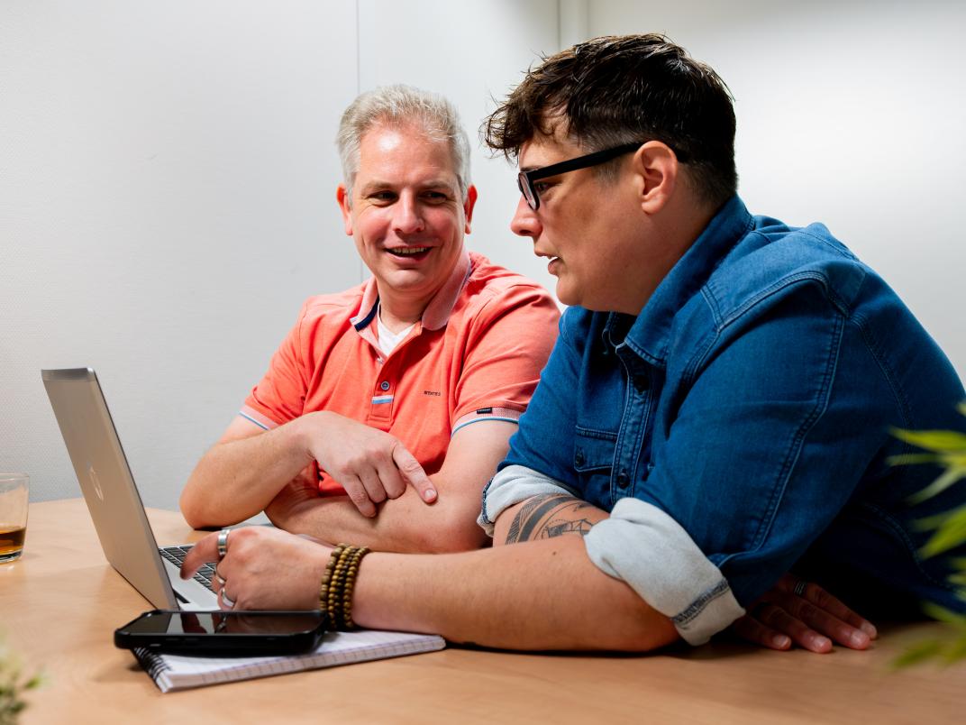 2 personen zitten aan tafel achter een laptop en zijn met elkaar in gesprek