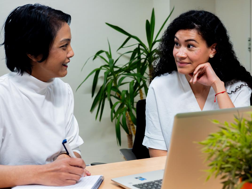 2 vrouwen kijken elkaar aan. 1 heeft een pen vast. Ze zitten achter een laptop, aan tafel. Er staat een plant voor de laptop en achter de 2 vrouwen.