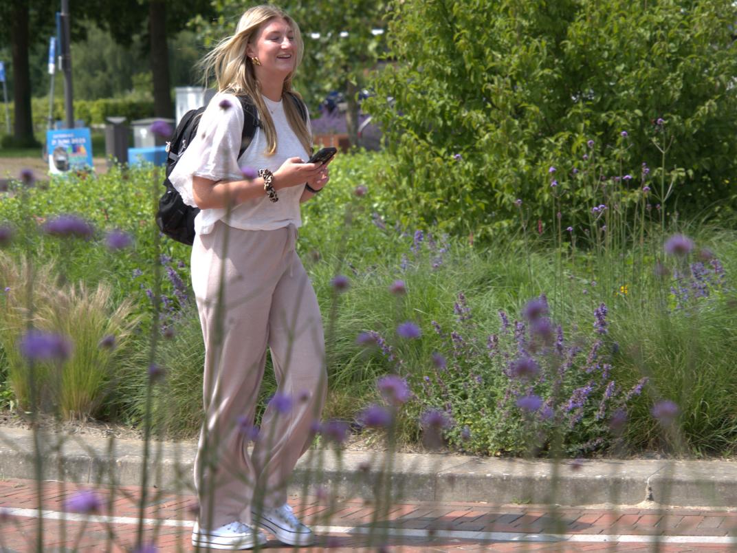 Vrouw loopt over straat, ze heeft een rugzak op en haar telefoon vast. Bloemen op de voorgrond, struiken op de achtergrond.