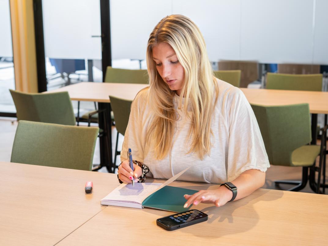 Vrouw zit aan tafel en schrijft in een boekje en tikt op de rekenmachine