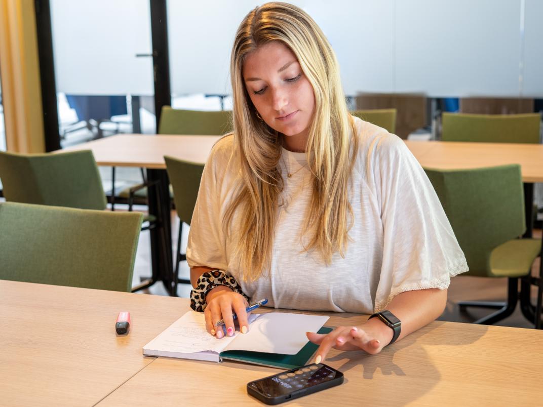 Vrouw zit aan tafel en tikt op haar rekenmachine. Er ligt een notitieboekje op tafel en ze heeft een pen vast.