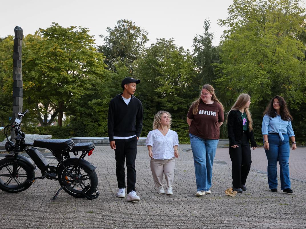 5 jongeren lopen naast elkaar buiten op straat, links staat een fatbike geparkeerd