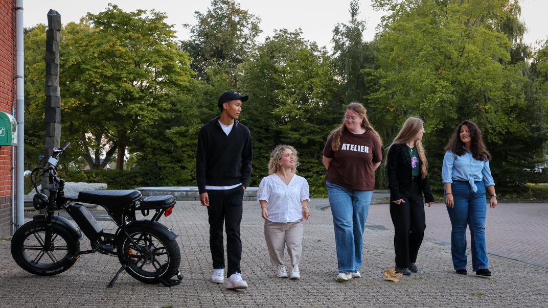5 jongeren lopen naast elkaar buiten op straat, links staat een fatbike geparkeerd